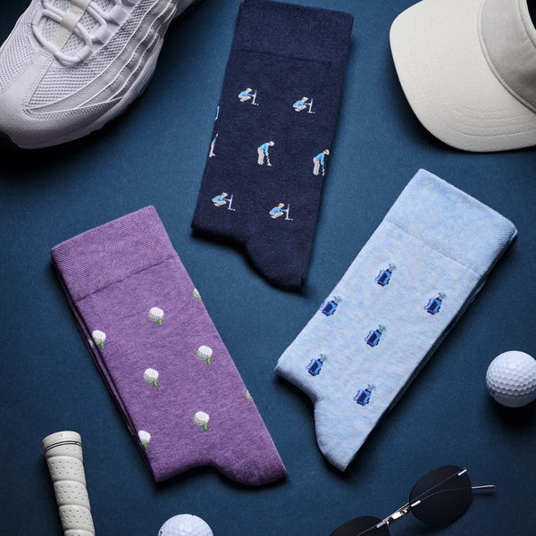 Three pairs of socks from The Caddie's Gift Box featuring golfer, golf ball, and golf bag patterns in purple, navy, and blue.