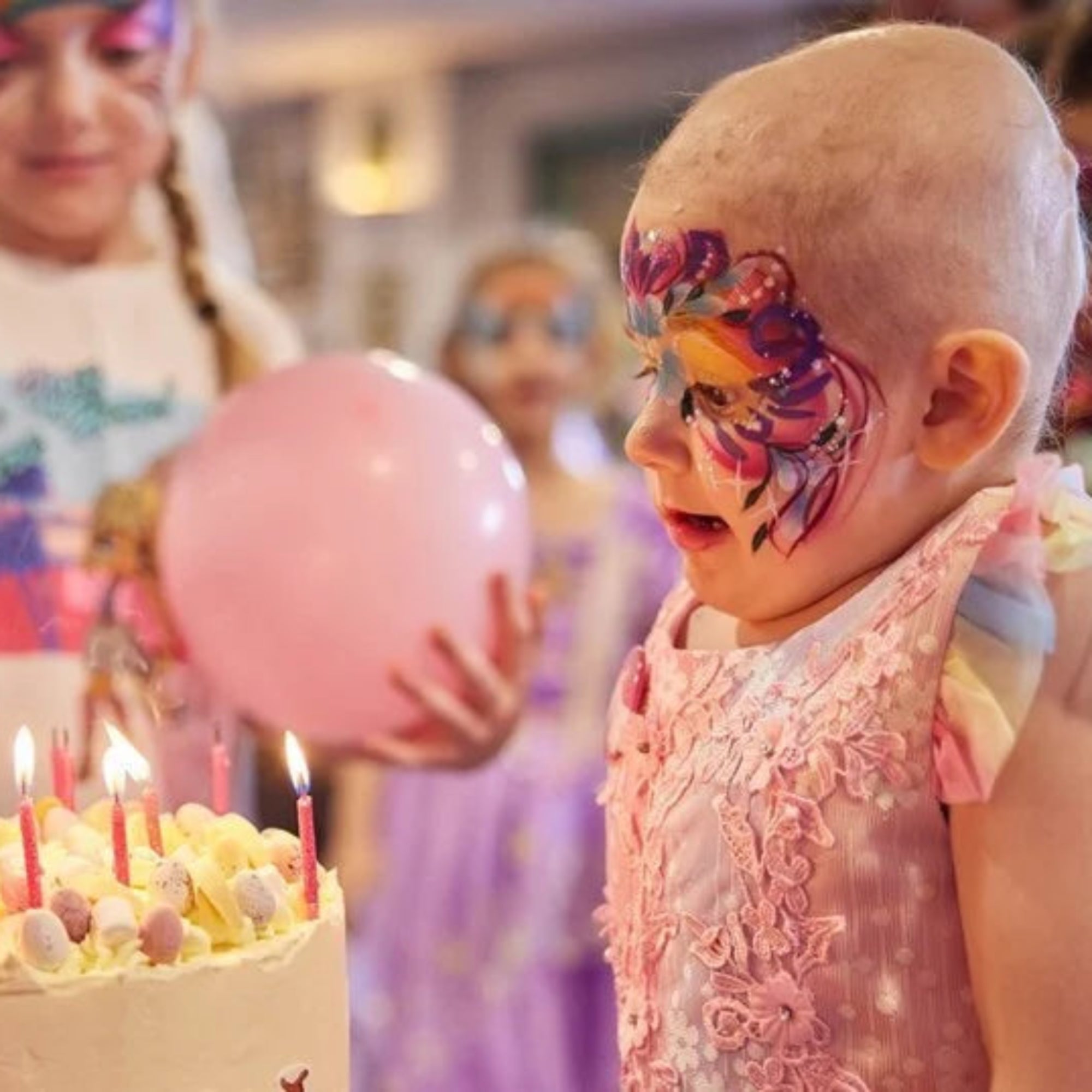 A baby in a pink dress with colorful face paint stands beside a birthday cake with lit candles and a pink balloon, surrounded by party guests in a festive indoor setting.