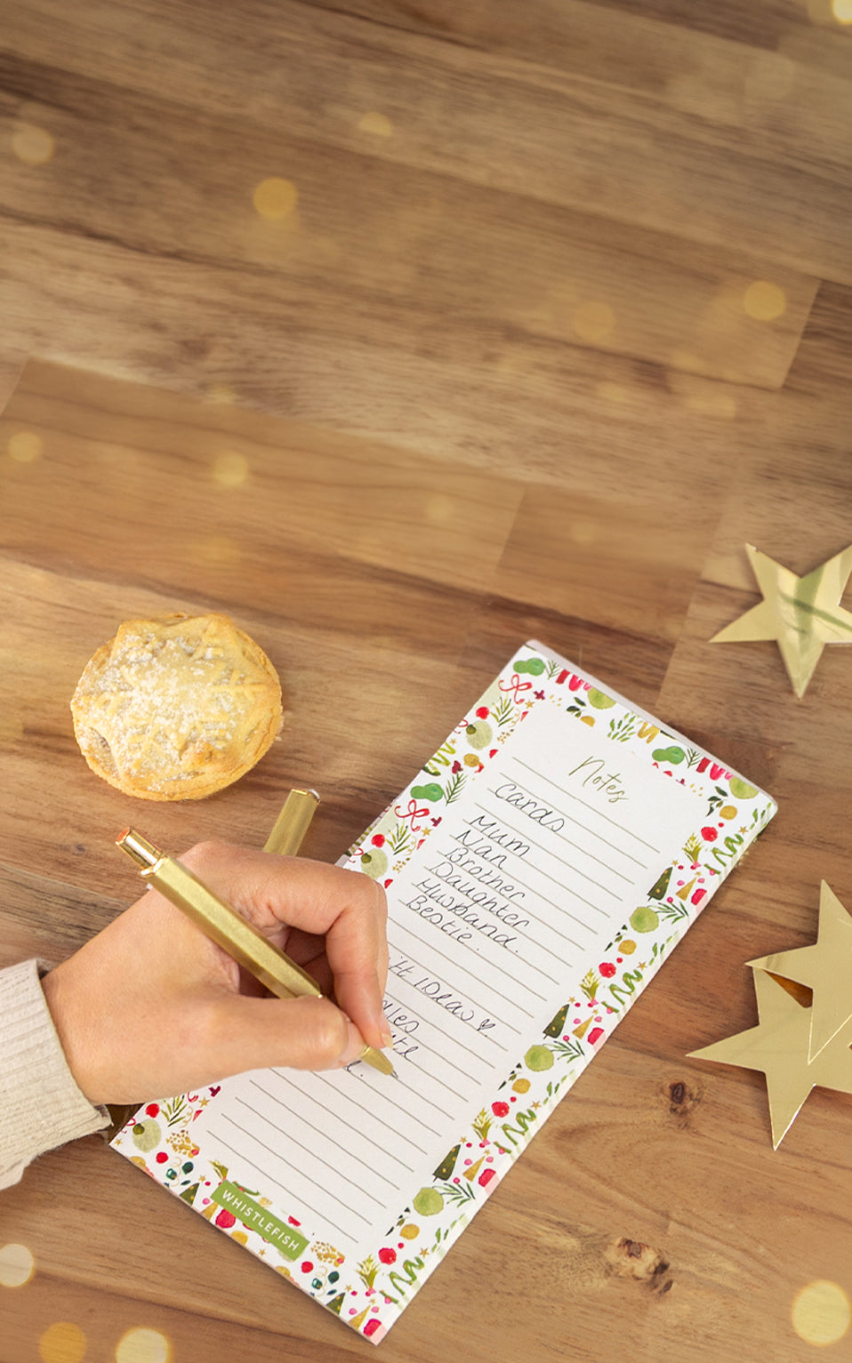 A hand holding a gold pen writes in a decorative holiday recipe notebook on a wooden table, with a pastry on a napkin and gold star decorations nearby.
