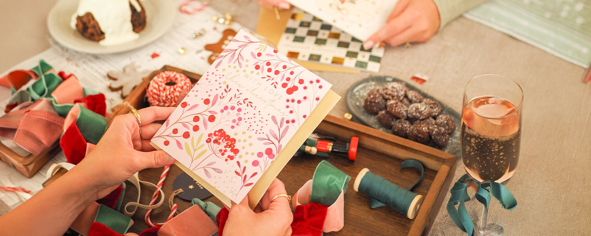 A person holds a decorated pink notebook or card with floral and berry patterns, surrounded by craft supplies, wrapped gifts, chocolate-covered treats, and a glass of rosé wine on a table.