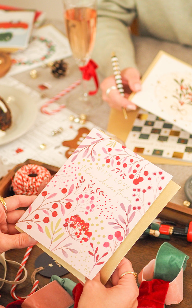 A person holds a decorated pink notebook or card with floral and berry patterns, surrounded by craft supplies, wrapped gifts, chocolate-covered treats, and a glass of rosé wine on a table.