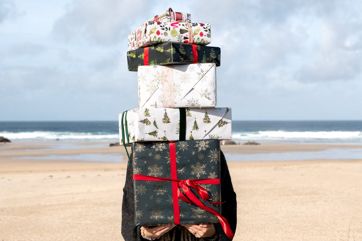 A stack of four wrapped Christmas gifts tied with red ribbons stands on a beach with the ocean and cloudy sky in the background.
