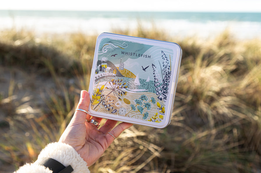 A notelet tin with a drawing of a coastal scene on the front being held in the sand dunes with the ocean behind
