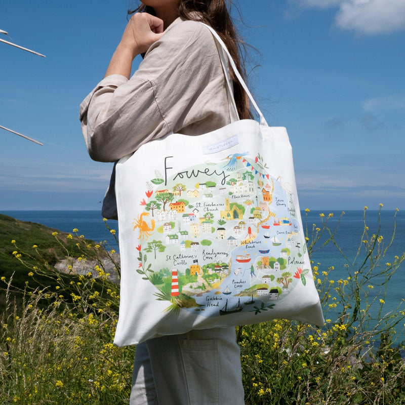 White canvas tote bag featuring a colourful illustrated map of Fowey, worn over the shoulder against a coastal backdrop.