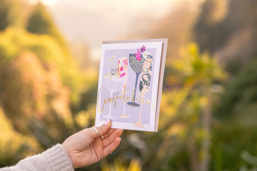 A luxury greetings card featuring champagne glasses being held up in the  late sunlight