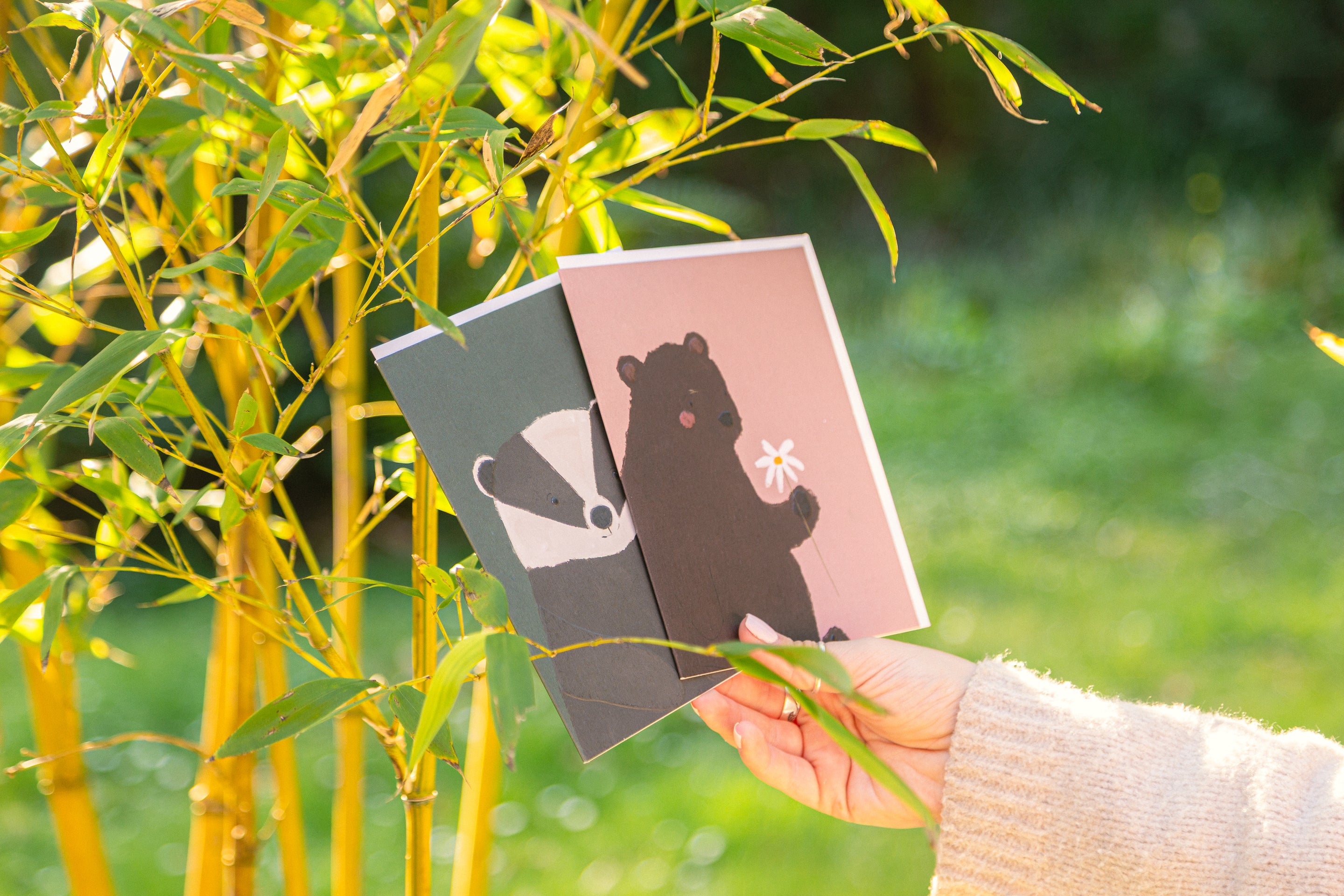 Two greetings cards featuring animals being held up against some bamboo in a garden