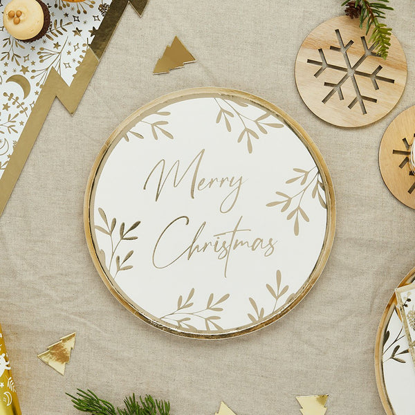 Round white paper plate with gold Merry Christmas script and botanical leaf illustrations on a linen tablecloth.