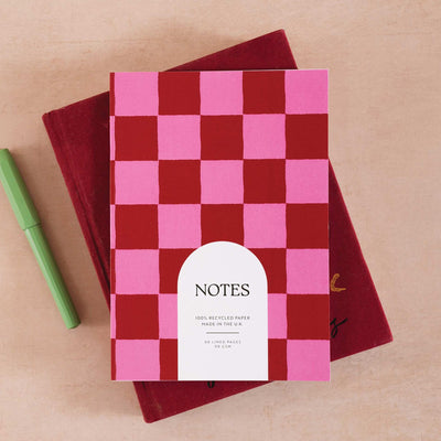 Burgundy and pink checked A5 notebook with a white arch label reading NOTES, resting on a dark red book with a green pen.