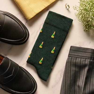 Dark green socks featuring a repeating pattern of small champagne bottles, laid out with black loafers and grey trousers.