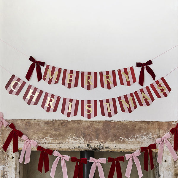 Stripey Christmas banner with gold lettering and deep red velvet bows, hung in two rows against a white wall.
