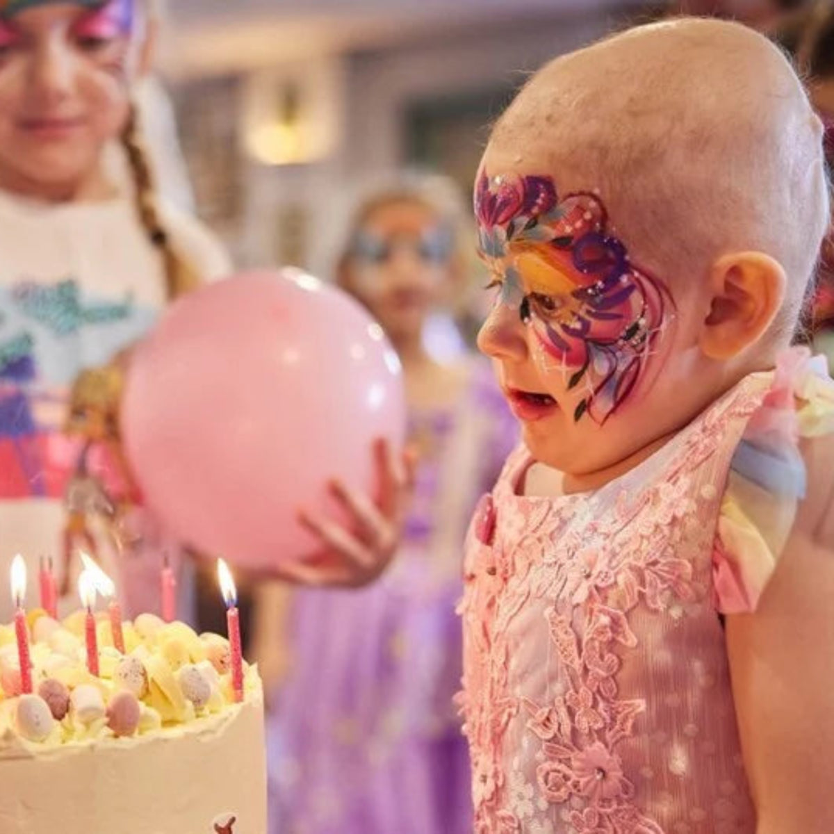 A baby in a pink dress with colorful face paint stands beside a birthday cake with lit candles and a pink balloon, surrounded by party guests in a festive indoor setting.