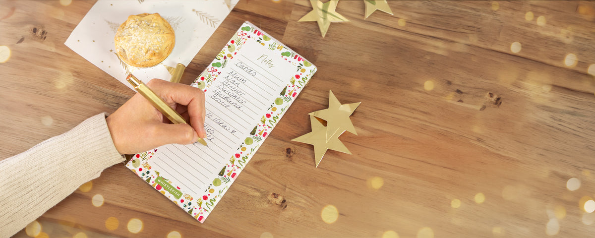 A hand holding a gold pen writes in a decorative holiday recipe notebook on a wooden table, with a pastry on a napkin and gold star decorations nearby.