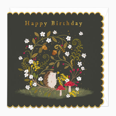Hedgehog Hideaway Birthday Card with a cute hedgehog, red toadstools, and white flowers on a dark background with gold edges.