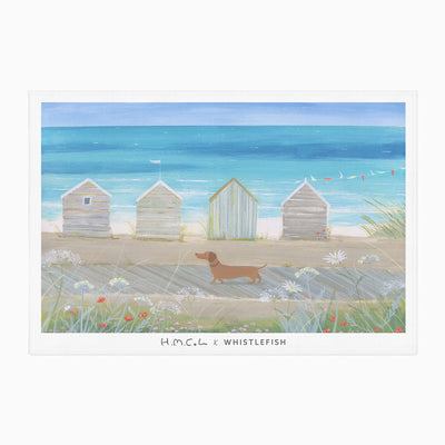 Sausage on The Shore tea towel featuring a dachshund walking past beach huts on a sandy path by the blue sea.