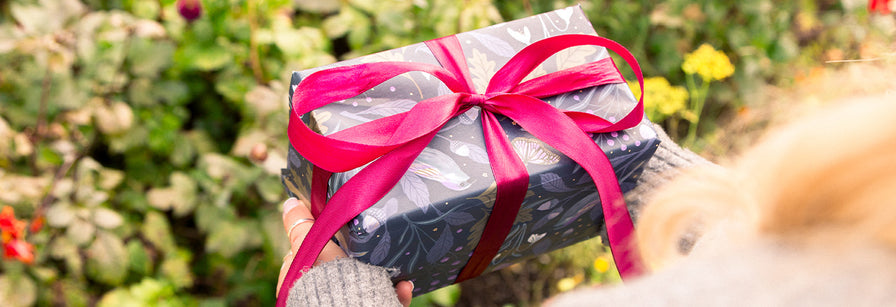 A beautifully wrapped Christmas present featuring dark floral wrapping paper and a bright pink ribbon, held outdoors in a garden.