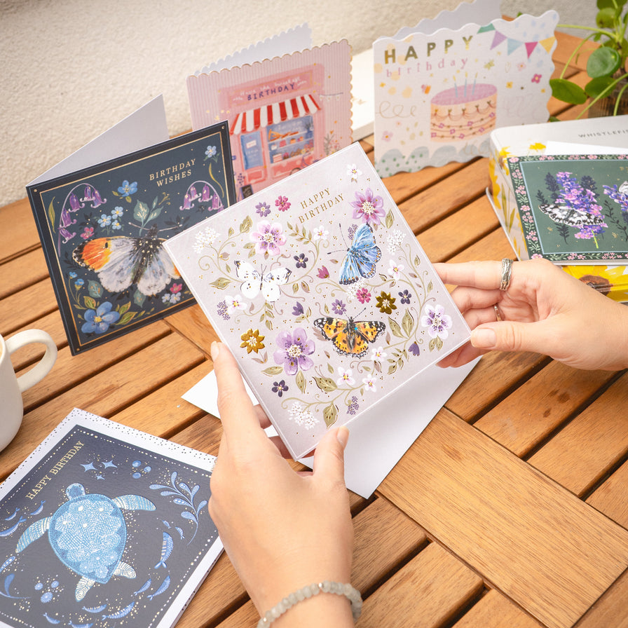 A person's hands hold a square white greeting card decorated with colorful butterflies, flowers, and a gold sunflower, with 'Happy Birthday' printed on it. Other illustrated greeting cards are arranged on a wooden table in a warm, natural setting.