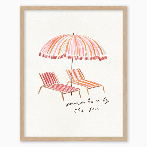 Somewhere by the Sea pink framed print featuring a striped beach umbrella and two sun loungers in a light wood frame.
