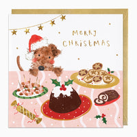 Puppy At The Table Christmas card featuring a dog in a Santa hat, mince pies, pudding, and gold stars on a pink tablecloth.