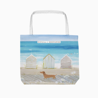 Sausage on The Shore tote bag featuring a dachshund walking past beach huts on a sandy path by the blue sea.